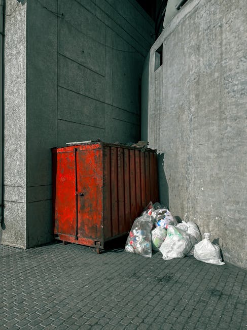 A large, industrial red metal skip with a weathered finish is positioned against a tall, dark green wall and a concrete wall in an urban setting. The skip is filled with various waste materials and is