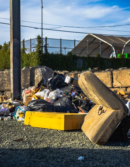 A waste collection technician wearing a high-visibility yellow and red uniform is standing at the rear of a large red rubbish truck on a paved roadside area, manually emptying a black plastic bag into